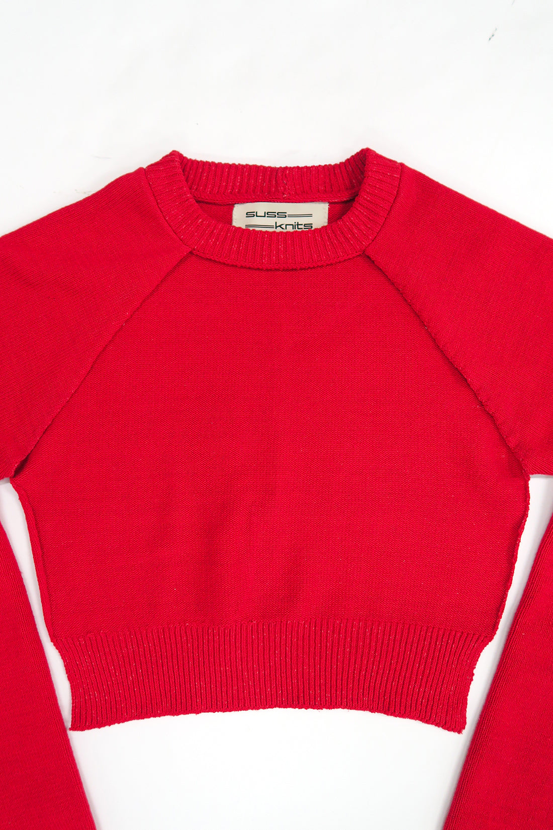 The Suss Knits Open Raglan knit in bright red cotton with raglan sleeves, laid flat on a white background. The brand label inside the collar reads 'SUSS KNITS'