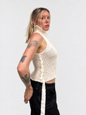 A woman modeling the suss knits chunky cream cotton sleeveless turtleneck, side profile view to see the silk ribbon detail