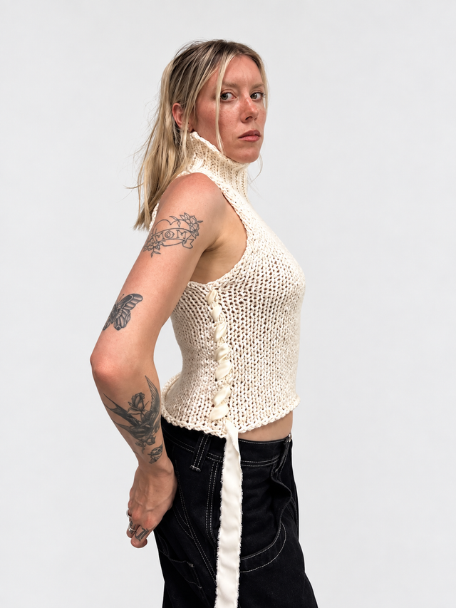 A woman modeling the suss knits chunky cream cotton sleeveless turtleneck, side profile view to see the silk ribbon detail
