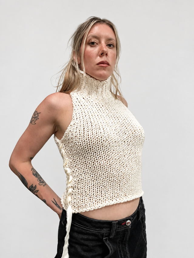 A woman modeling the Suss Knits chunky cream cotton sleeveless knit turtleneck with silk ribbon side detail stitching