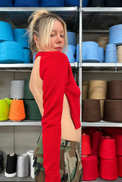 A woman with blonde hair in a messy bun stands in a studio or workshop setting with shelves of colorful yarn spools behind her. She is wearing the Suss Open Raglan in bright red cotton with long-sleeves and a dramatic asymmetrical cutout design on the back and side, paired with camouflage cargo pants.